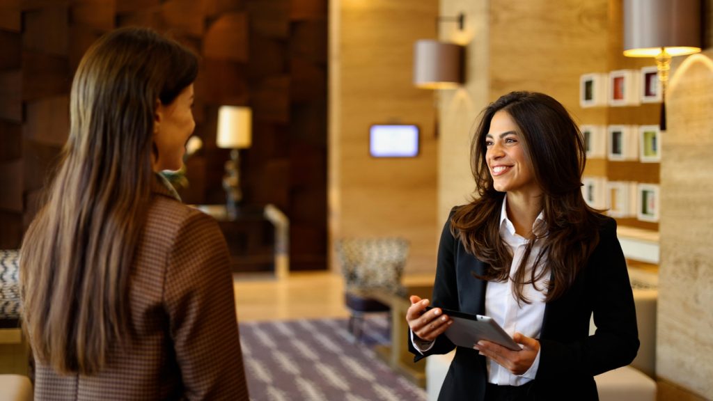 Ein lächelnder Hotelmitarbeiter, der ein Tablet bedient, während er sich mit einer weiblichen Gastin in der Lobby eines modernen Hotels unterhält