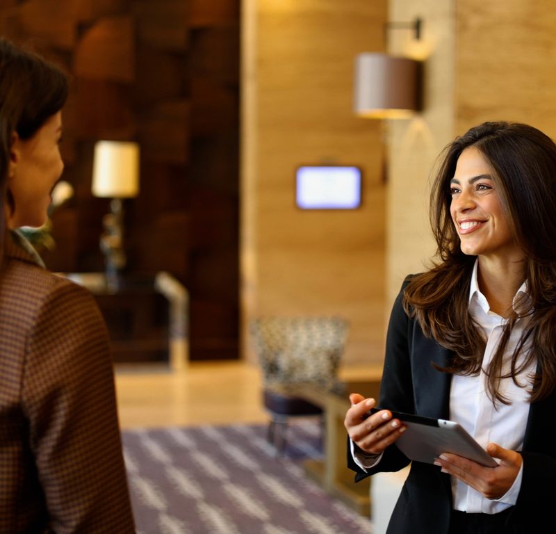 Ein lächelnder Hotelmitarbeiter, der ein Tablet bedient, während er sich mit einer weiblichen Gastin in der Lobby eines modernen Hotels unterhält